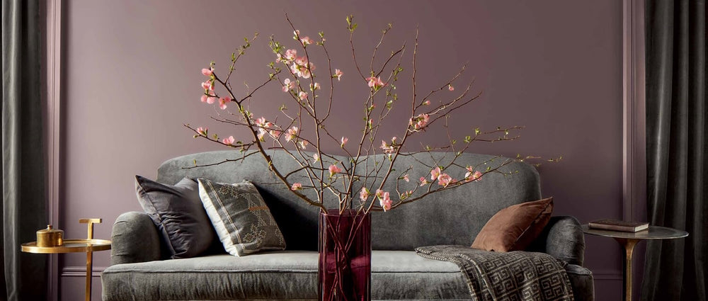 A purple-painted living room features a gray couch, gold side tables, and a glass-topped gold coffee table topped with a red vase filled with pink flowers.